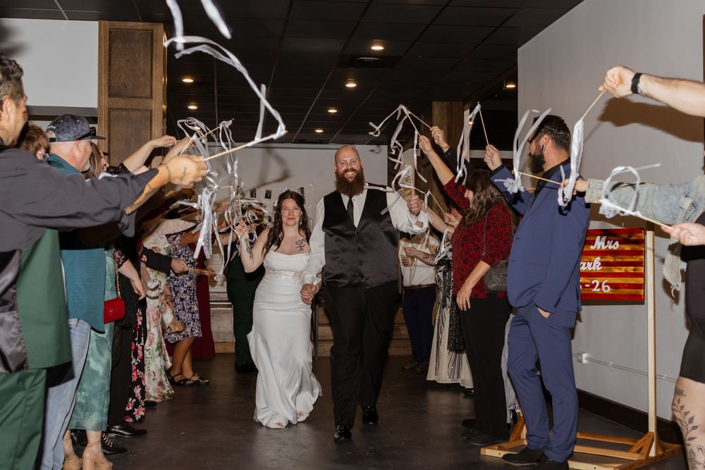 A bride and groom exit their wedding reception through a celebratory ribbon wand tunnel surrounded by cheering guests.