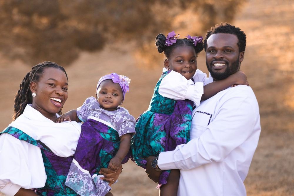 Happy Black family posing outdoors, parents holding two young daughters in matching purple and teal floral dresses.