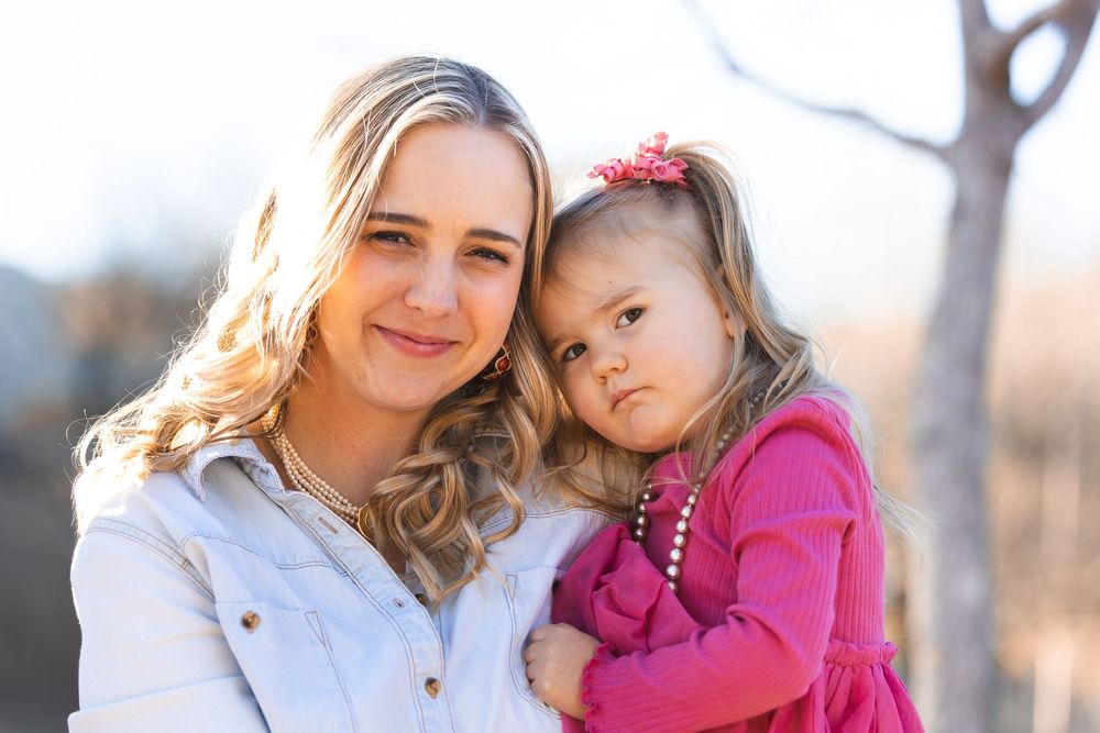 A smiling blonde woman holds a young toddler girl in a pink dress and bow outdoors in warm sunlight.