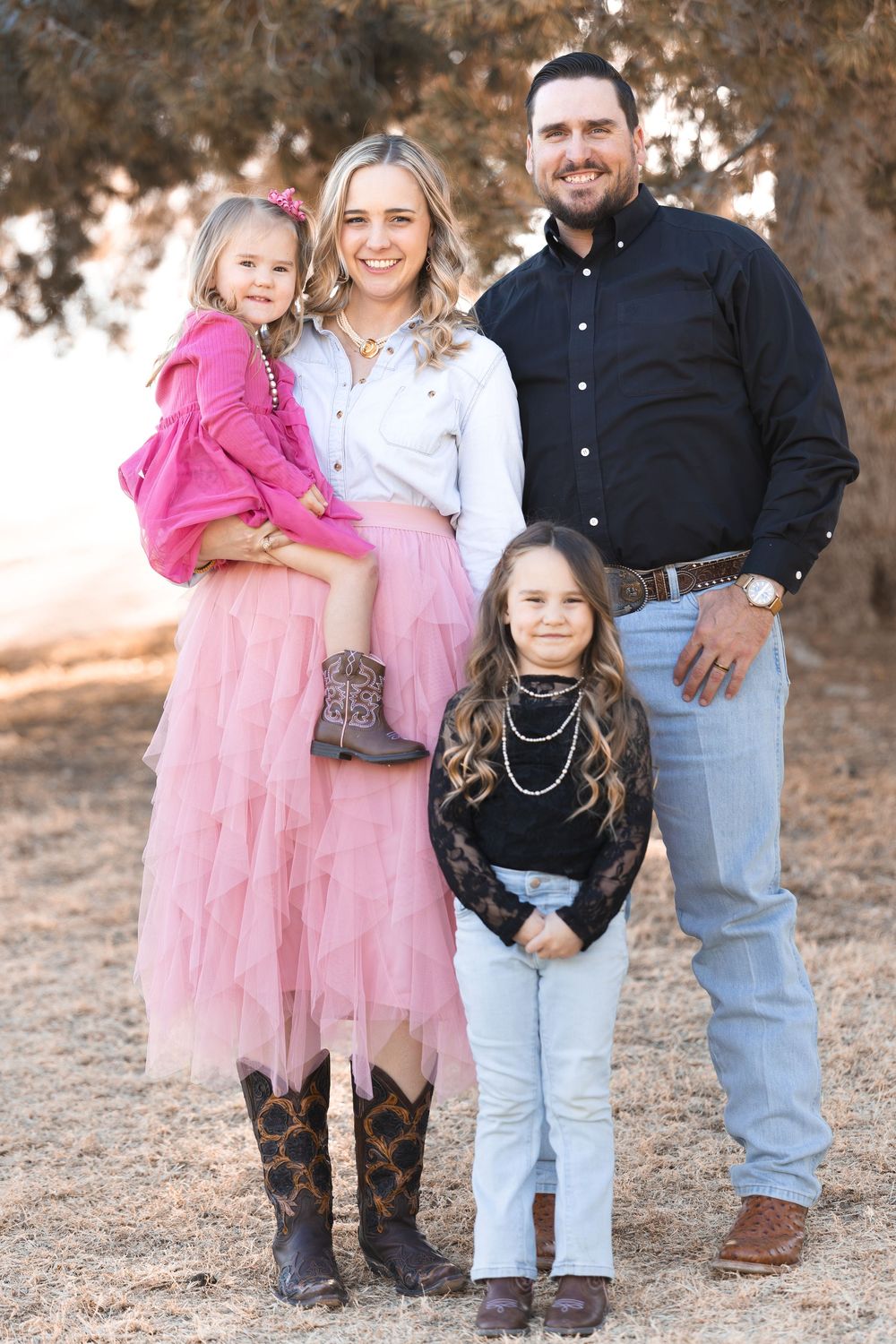 A smiling family of four poses outdoors, featuring two girls in pink outfits and cowboy boots with their parents.
