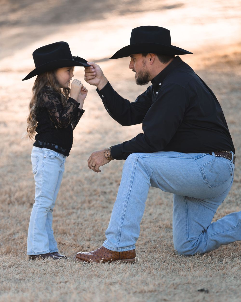 A man in a black cowboy hat kneels to adjust a young girl's matching black cowboy hat outdoors.