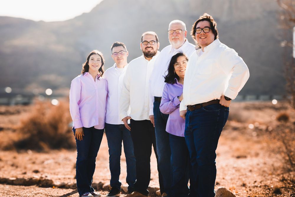 Six people smiling outdoors in a desert landscape with mountains, wearing casual white and purple tops for a family photo.