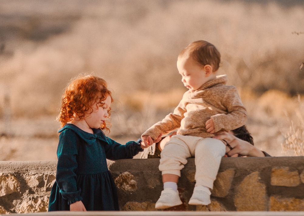 Two young children, a red-haired toddler girl in a teal dress and a baby boy, hold hands on a stone wall outdoors.