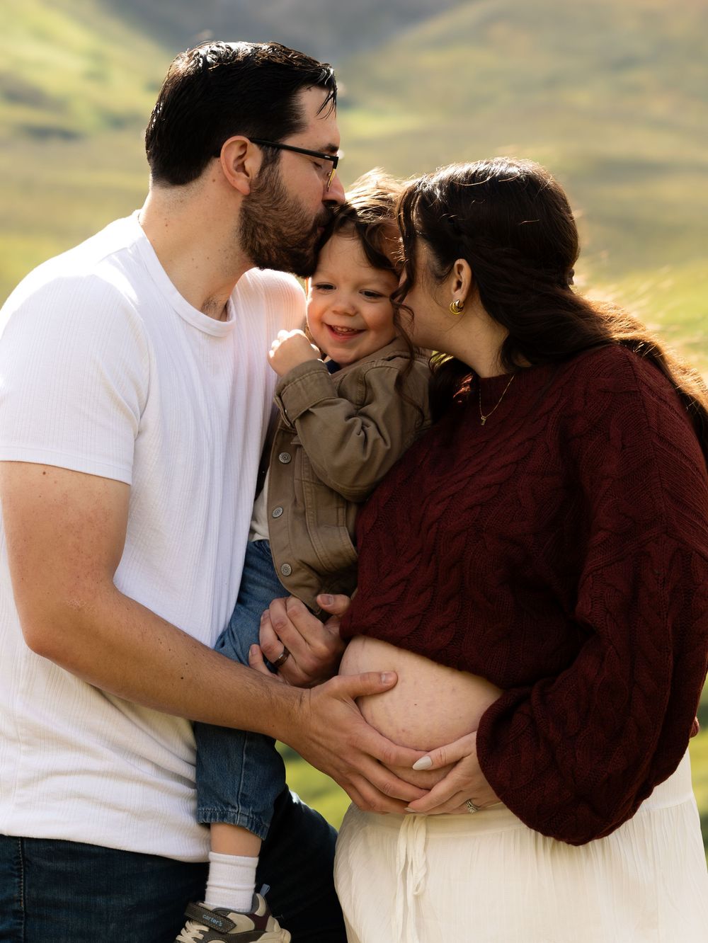 A happy family maternity photo with a man and pregnant woman kissing their smiling toddler outdoors.