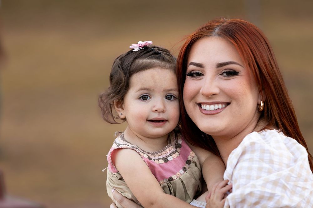 A smiling red-haired woman holds a cute toddler girl with a pink flower clip outdoors.