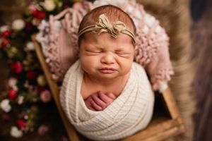 Newborn baby girl wrapped in white swaddle with gold bow headband sleeping in wooden crate with floral backdrop.
