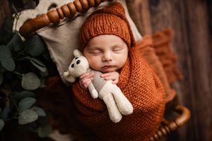Sleeping newborn baby wrapped in rust orange knit blanket wearing bear hat, holding stuffed animal toy in cozy basket.