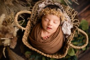 Sleeping newborn baby wrapped in brown swaddle wearing gold floral crown, nestled in wicker basket with greenery.