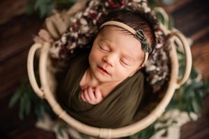 Newborn baby swaddled in olive green wrap sleeping peacefully in a wicker basket with floral accents.