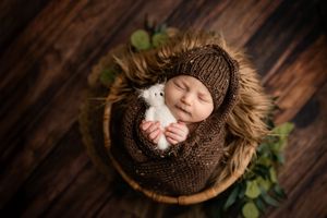 Newborn baby sleeping peacefully in a brown knit wrap inside a wooden bowl, holding a white stuffed bunny toy.