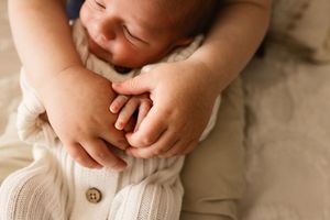 Close-up of a sleeping newborn baby with tiny hands clasped together, wearing a cream knit outfit on a beige blanket.
