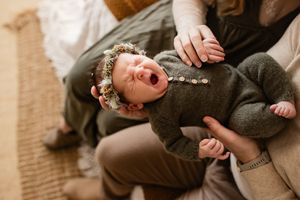 Crying newborn baby wearing floral crown in olive green romper held by parent during lifestyle newborn photography session.