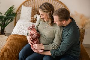 A couple sits on a bed smiling at their newborn baby wrapped in a pink swaddle in a cozy boho-styled room.