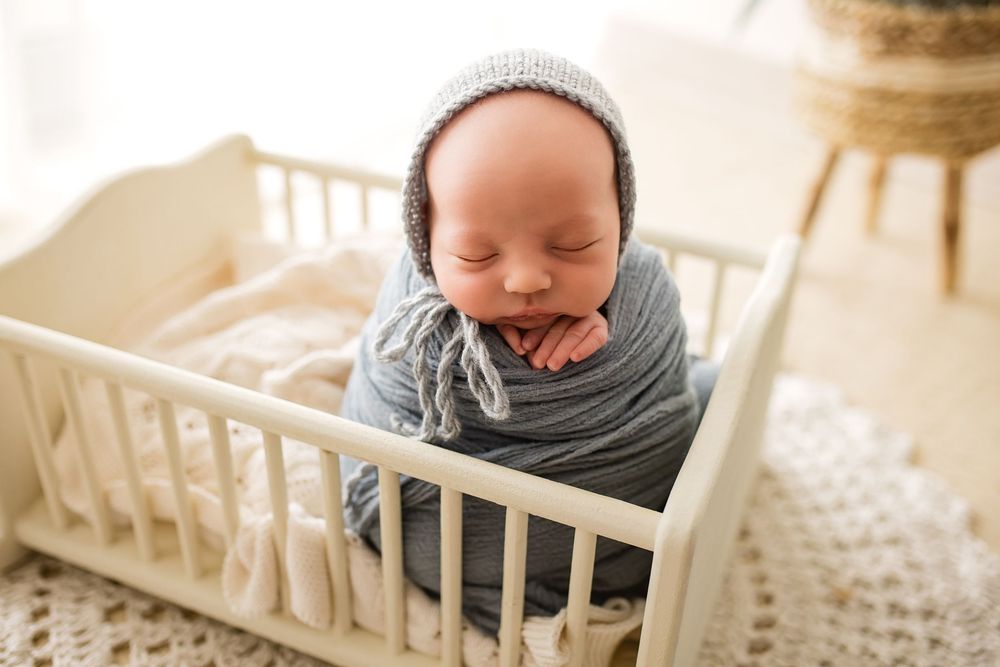 Sleeping newborn baby wrapped in gray swaddle wearing knit bonnet in a small white wooden crib.