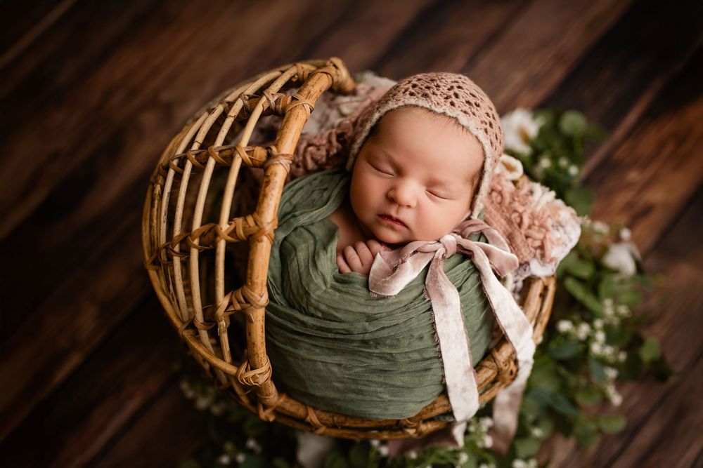 Sleeping newborn baby wrapped in green swaddle wearing pink bonnet resting in wicker basket with floral decor.