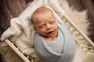 Newborn baby swaddled in gray wrap sleeping peacefully in a small white wooden bed with cream blanket.