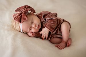 Sleeping newborn baby girl in dusty rose velvet outfit and bow headband on cream background.