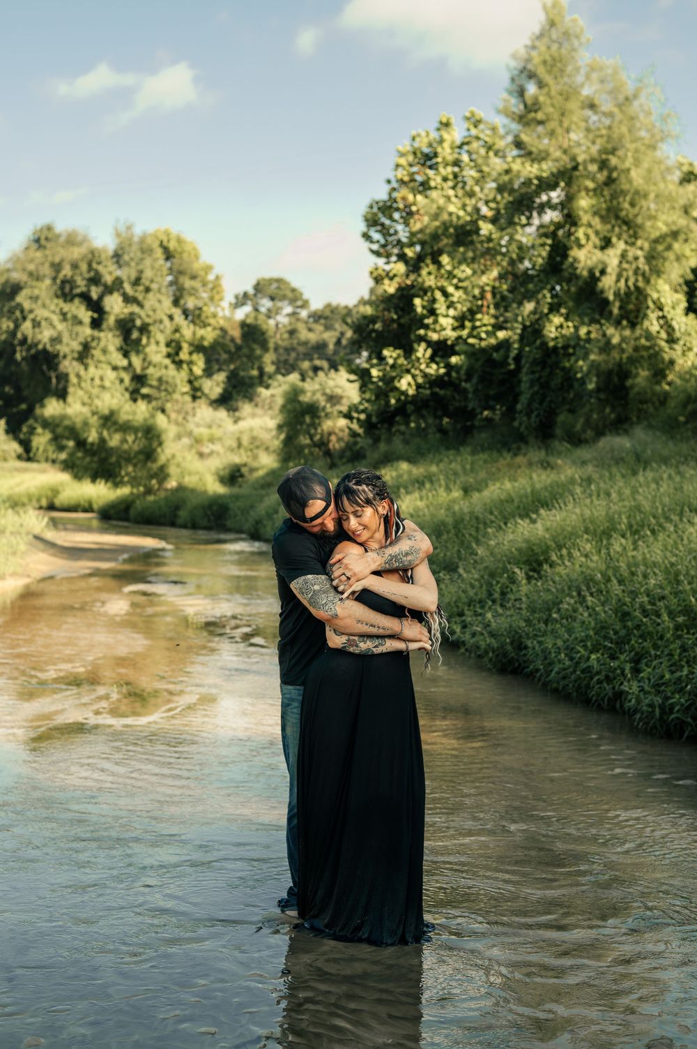 Couple embracing romantically in a shallow river surrounded by lush green trees on a sunny day.