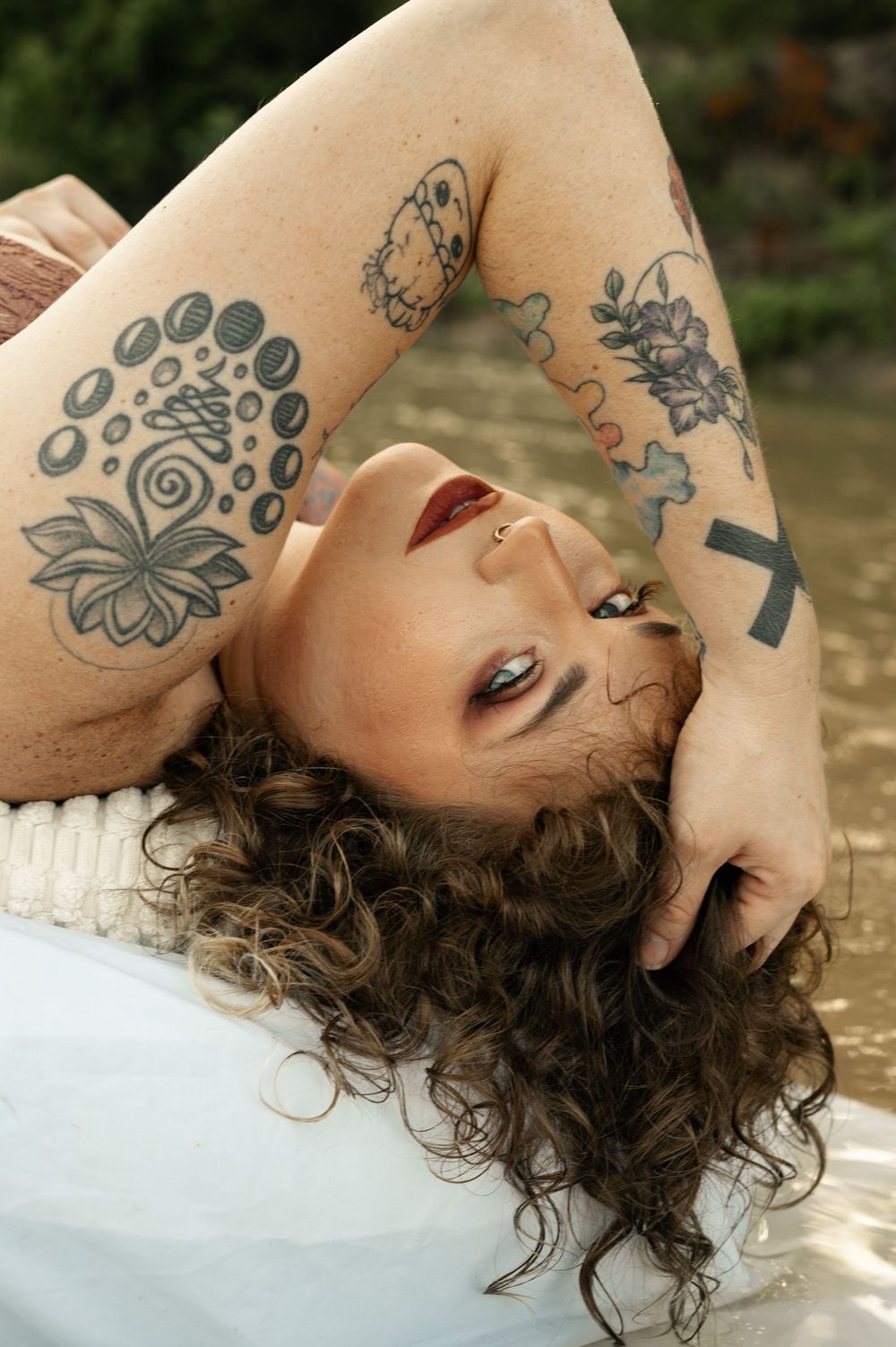 A tattooed woman with curly hair and red lips poses reclined, showcasing floral and abstract ink artwork on her arms.