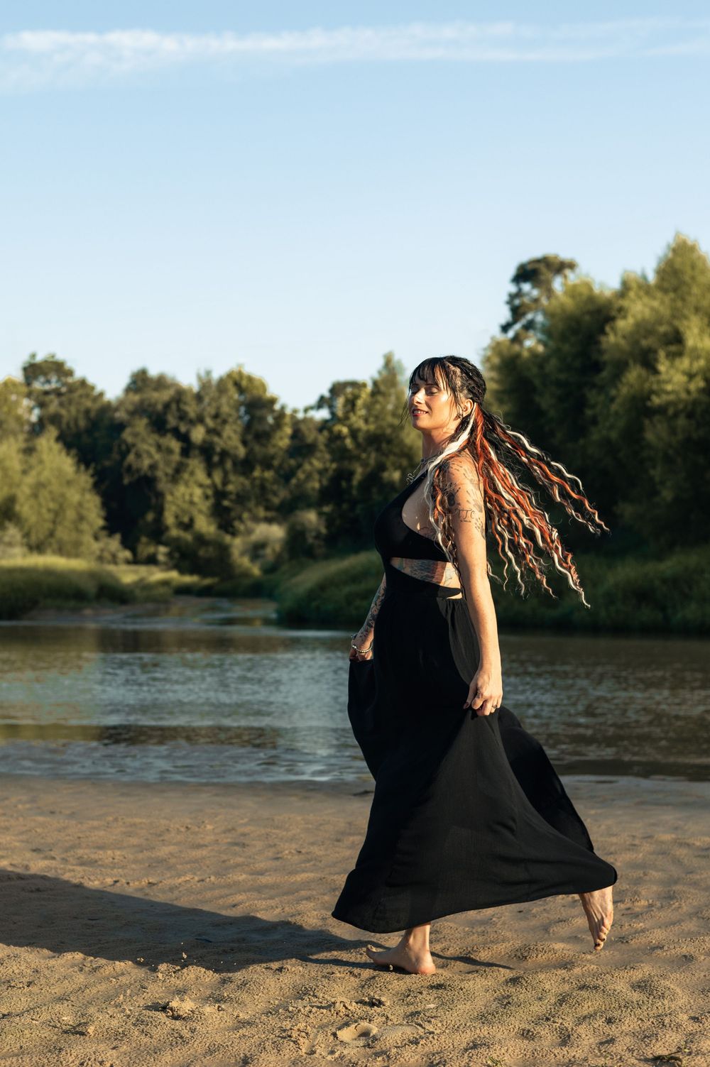 Woman in black maxi dress with colorful dreadlocks walking barefoot on sandy riverbank at golden hour.