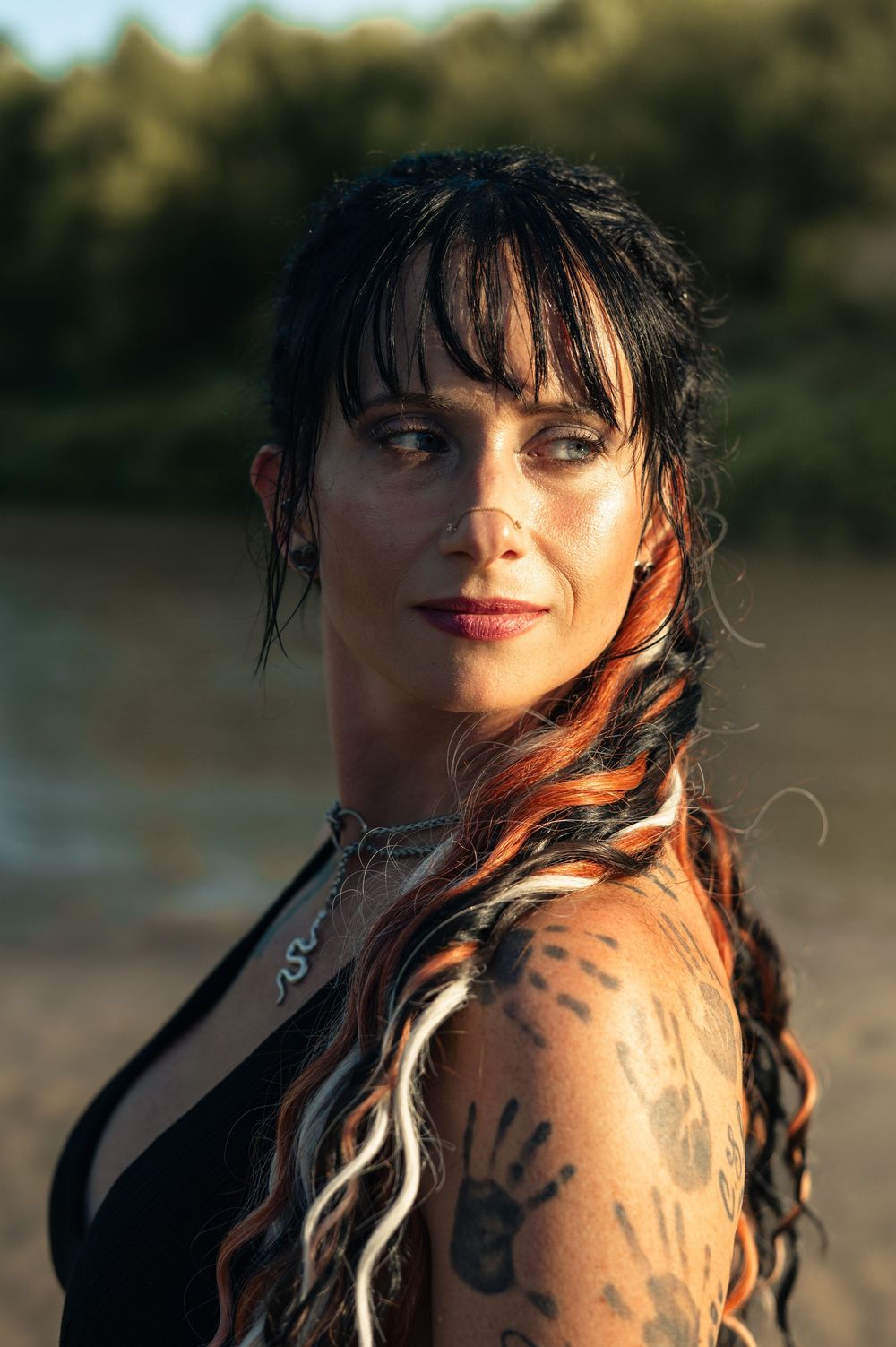 Woman with dark hair, colorful braids, and hand tattoos poses outdoors near water in golden hour light.