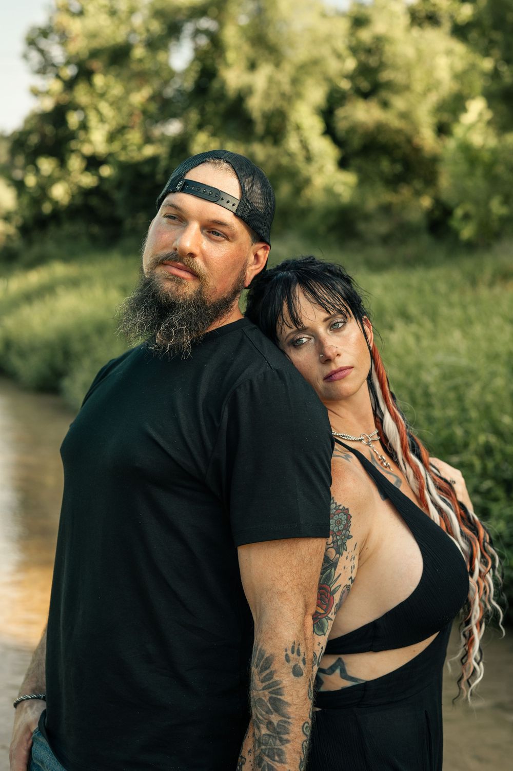 Tattooed couple in black outfits pose back-to-back near a creek surrounded by lush green trees.