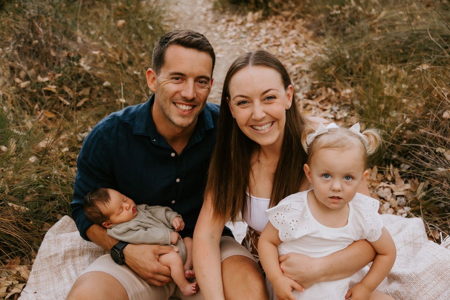Happy family of four sitting outdoors on a blanket, parents smiling with newborn baby and toddler girl.