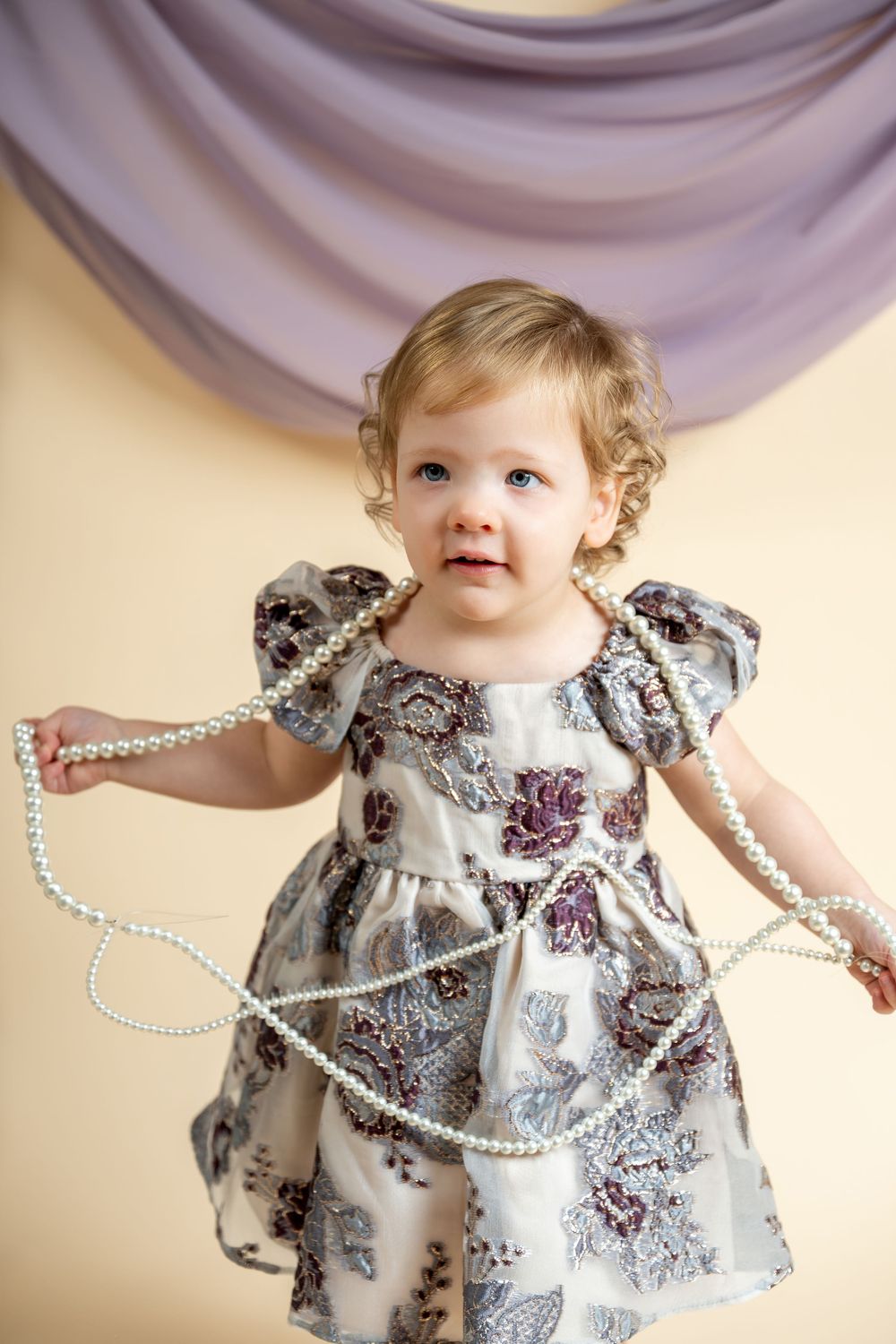 Toddler girl in floral dress playing with pearl necklace beads, posing in front of lavender draped backdrop.