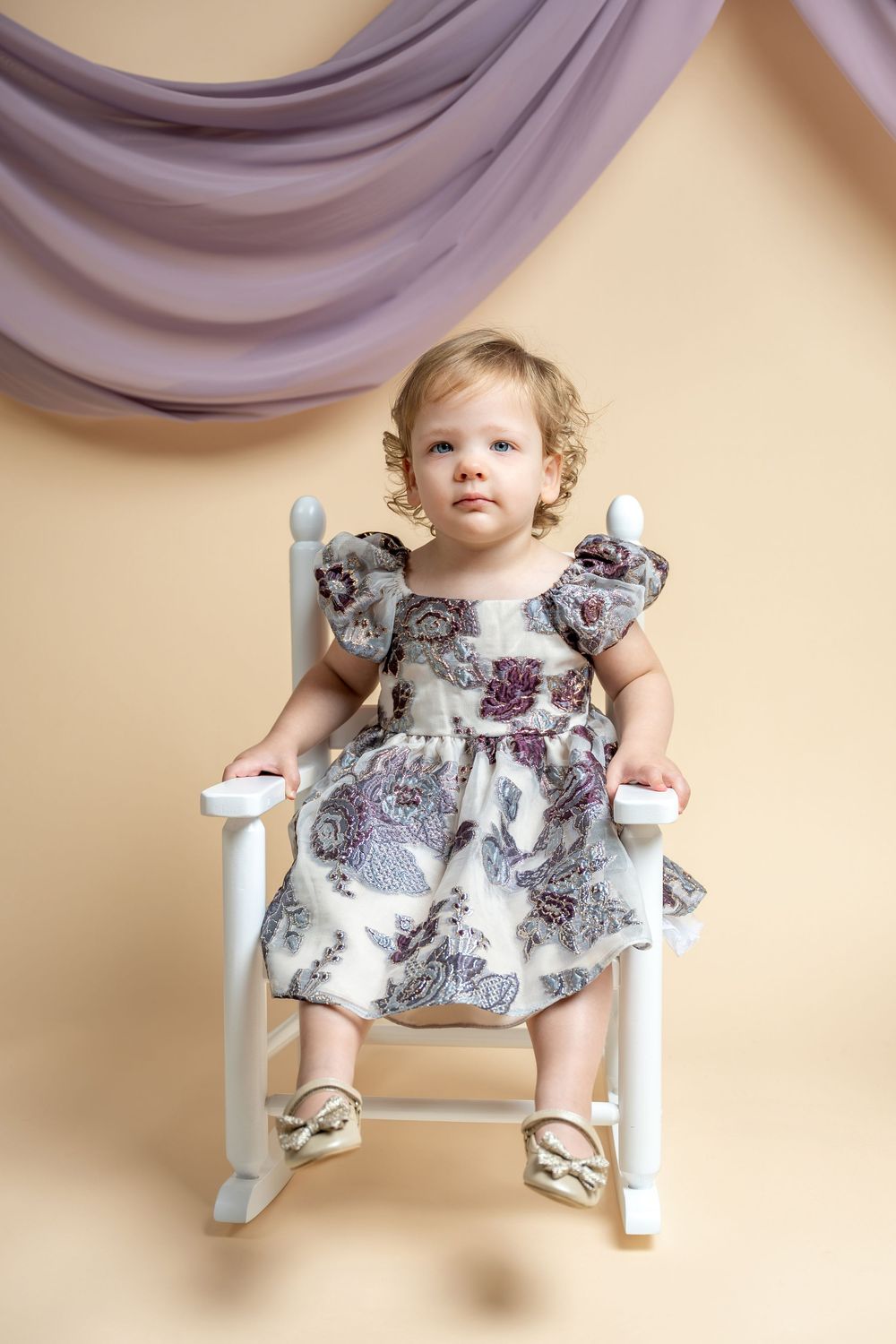 Toddler girl in floral dress sitting in white rocking chair against beige backdrop with purple draped fabric.