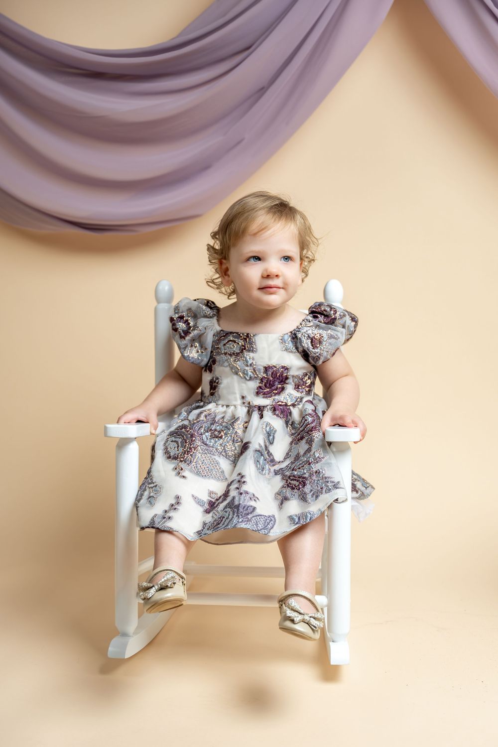 Adorable toddler girl in floral dress sitting in white rocking chair against beige backdrop with purple draping.