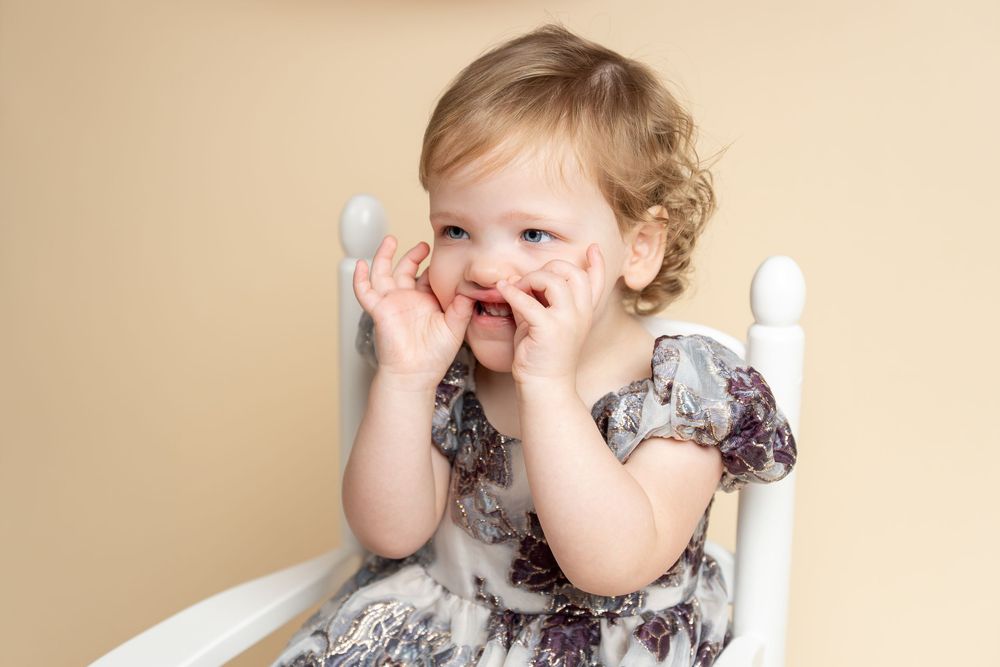 Adorable toddler girl in floral dress sitting in white chair, smiling with hands on cheeks against beige background.