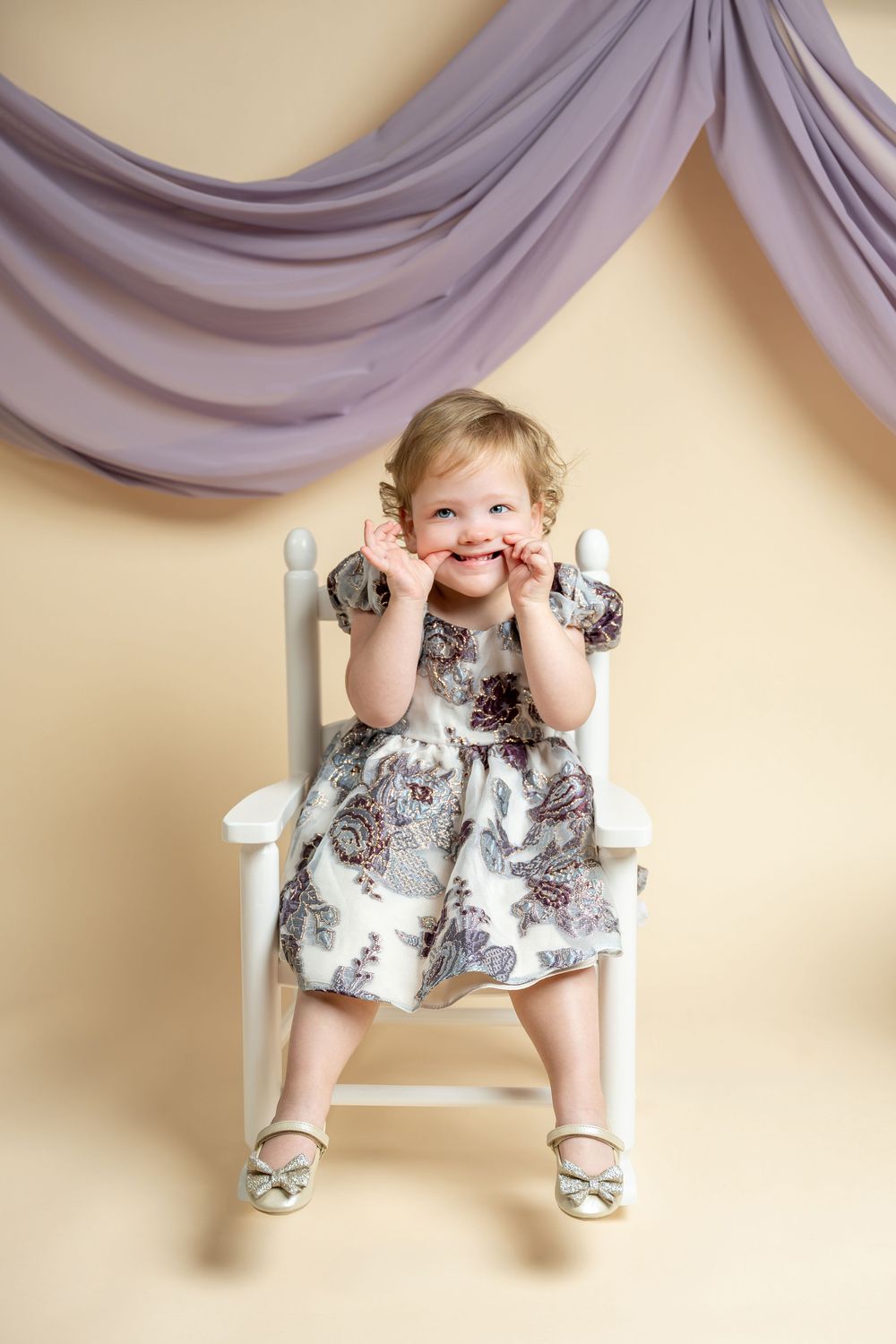 Adorable toddler girl in floral dress sitting on white chair, smiling with hands on cheeks, purple drape backdrop.
