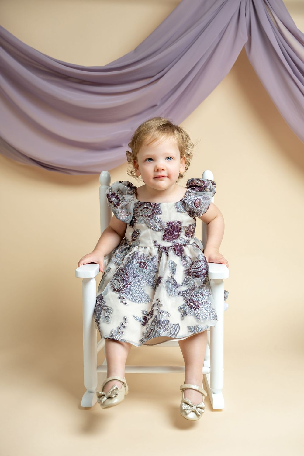 Toddler girl in floral dress sitting on white chair with purple drape backdrop.