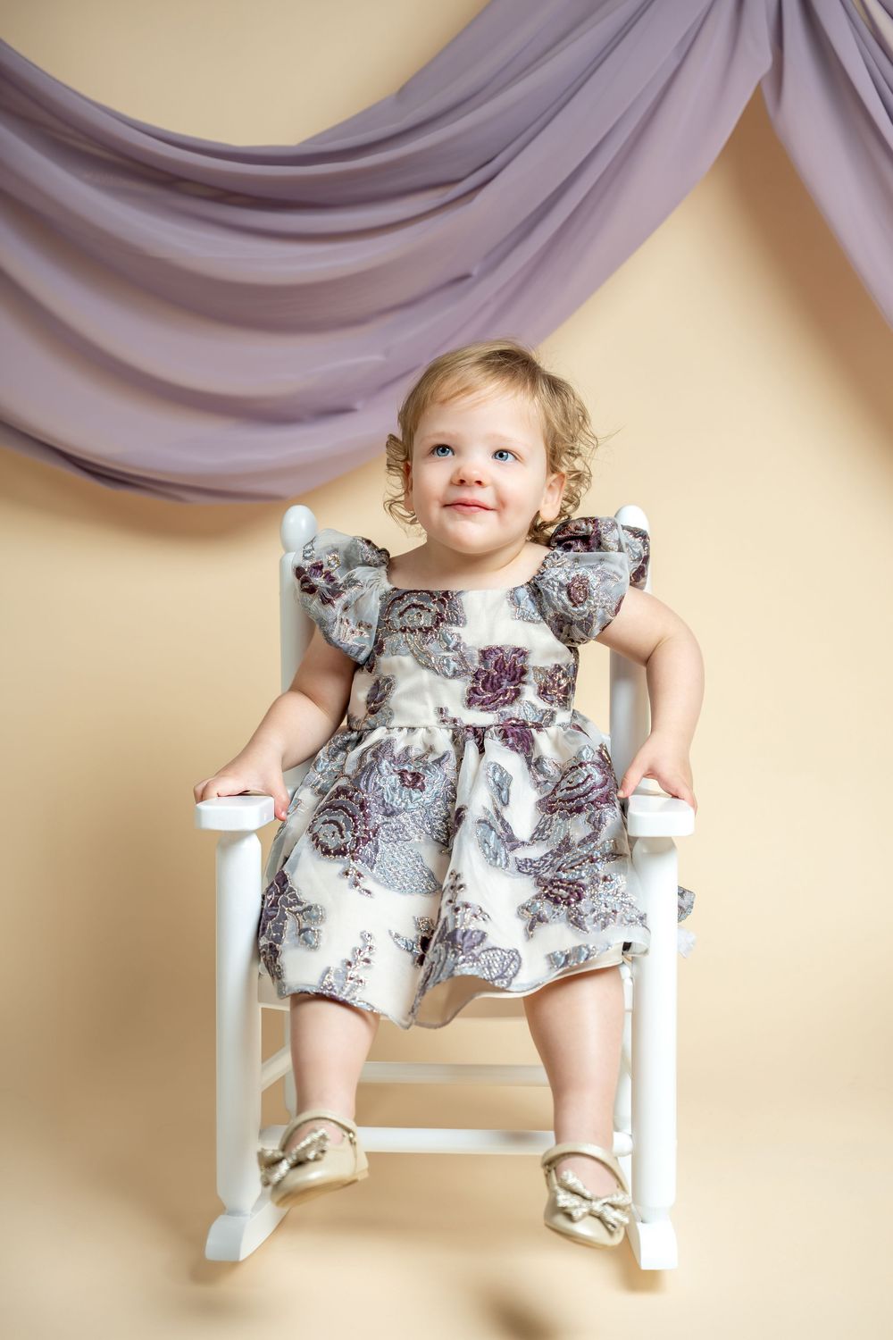 Adorable toddler girl in floral dress sitting on white rocking chair against beige backdrop with purple draping.