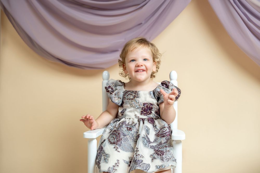Smiling toddler girl in floral dress sitting in white chair holding Easter eggs against beige backdrop.