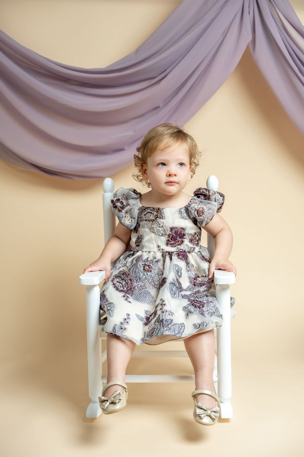 Adorable toddler girl in floral dress sitting in white rocking chair against beige backdrop with purple draping.