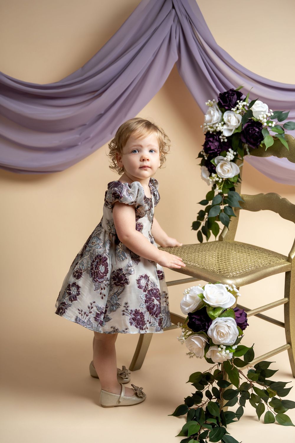 Toddler girl in floral purple dress standing by chair with white and purple roses, lavender drape backdrop.