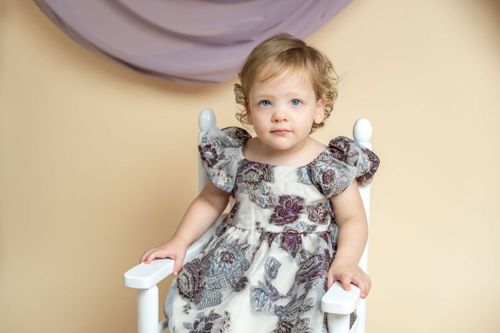 Adorable toddler girl in elegant floral dress posing in white chair against beige and purple backdrop.