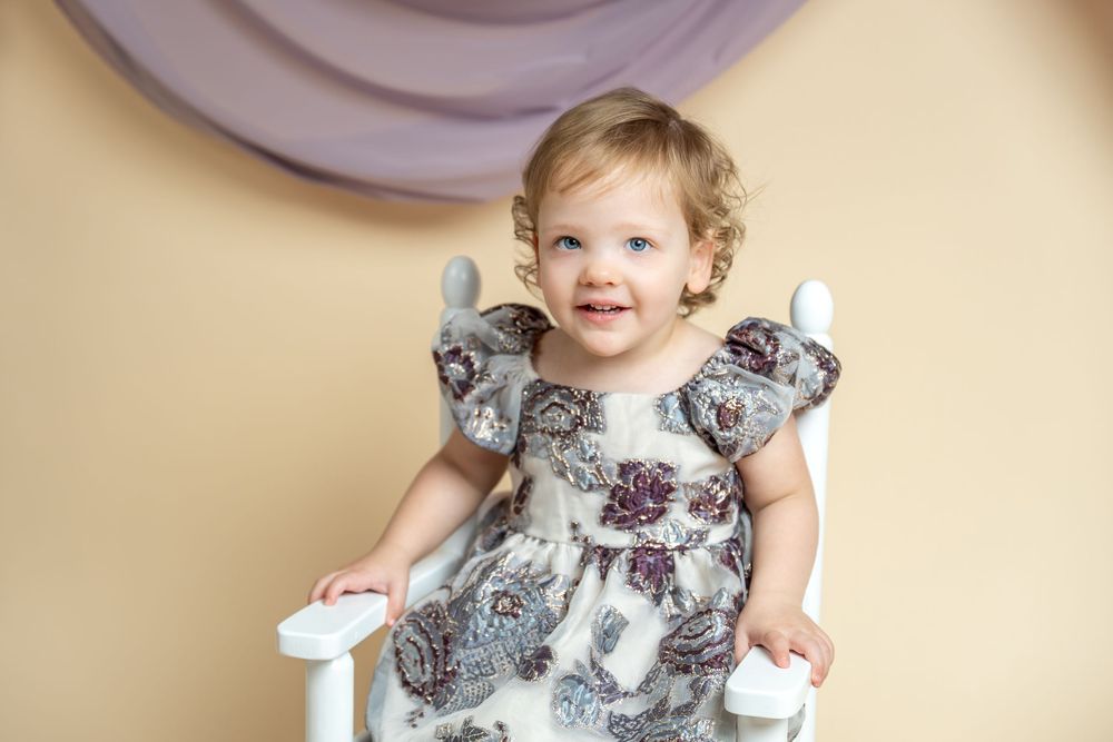 Smiling toddler girl in floral dress sitting on white chair against beige backdrop in professional studio portrait.