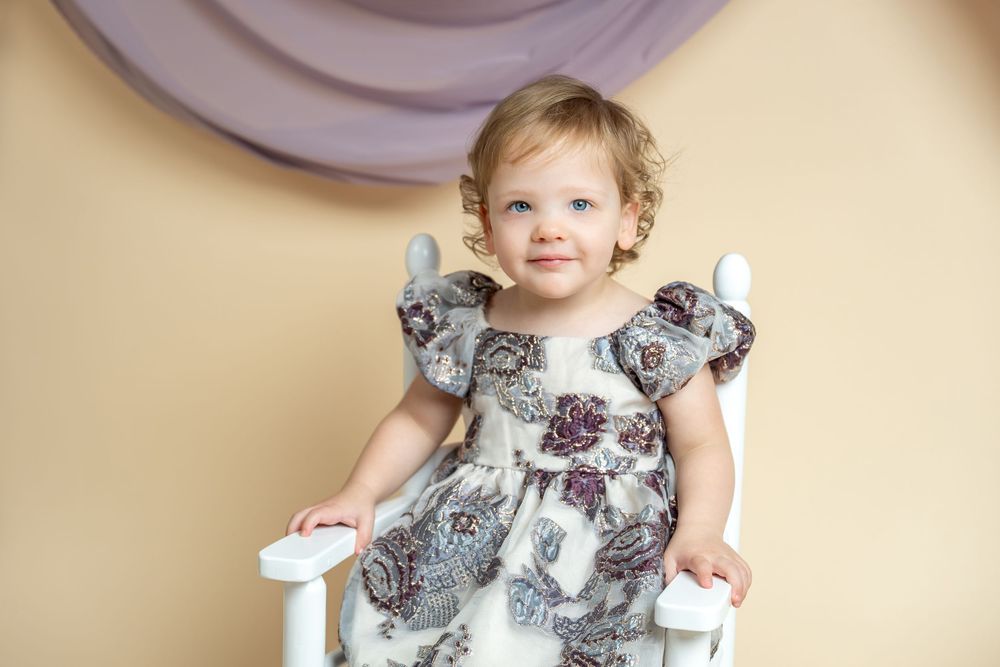 Adorable toddler girl in floral dress sitting on white chair against beige backdrop in professional portrait.