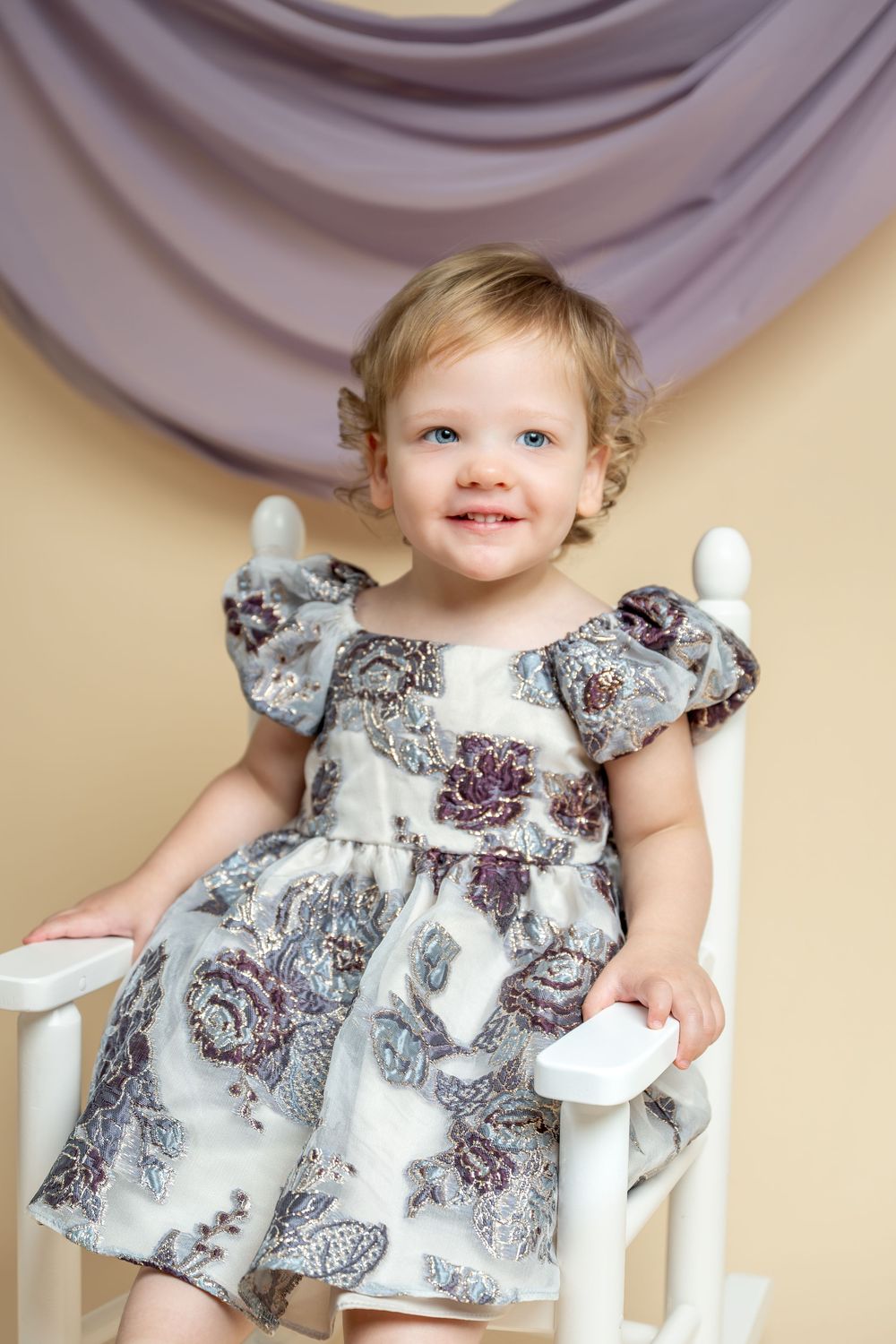 Smiling toddler girl in floral dress sitting on white rocking chair with purple drape background.