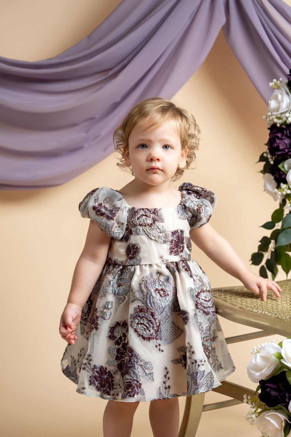 Toddler girl in floral dress poses in studio with purple drape and flowers.