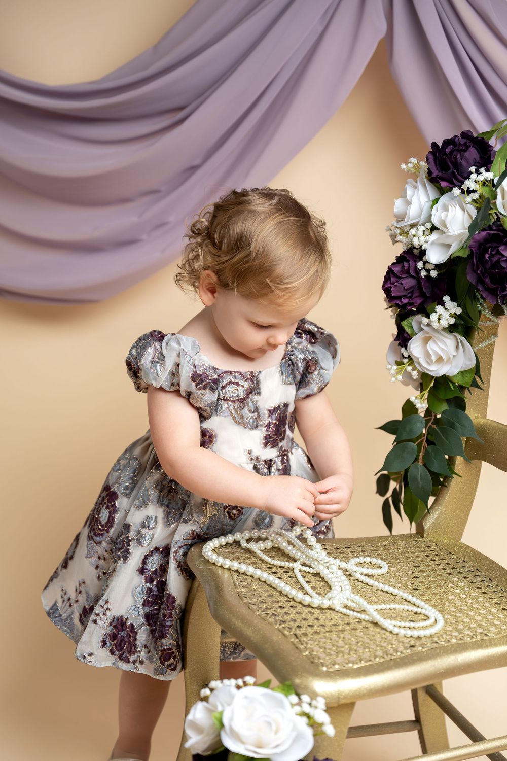 Toddler girl in floral dress playing with pearl necklace on gold chair, purple drapes and flowers in background.