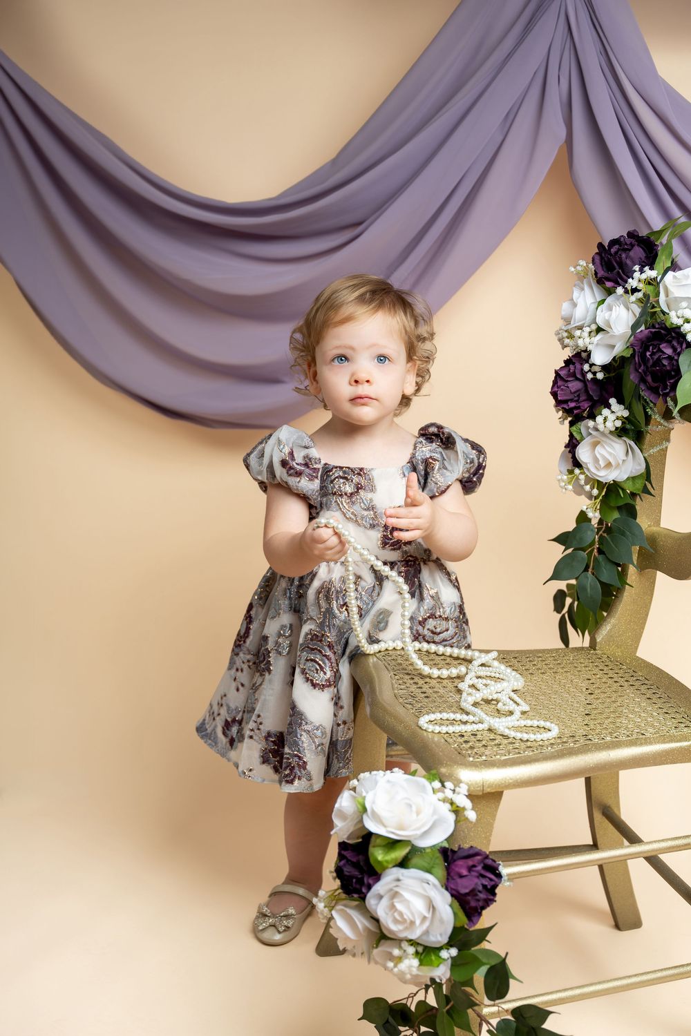 Toddler girl in floral dress holding pearls beside a chair with purple and white flowers.