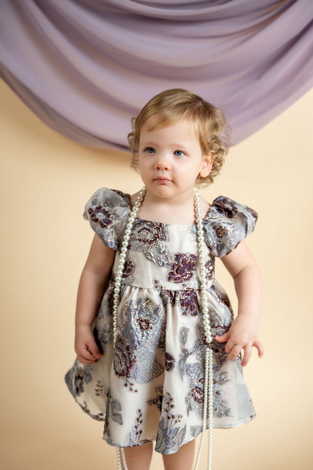 A toddler girl in a floral dress wearing pearl necklaces poses against a lavender draped background.