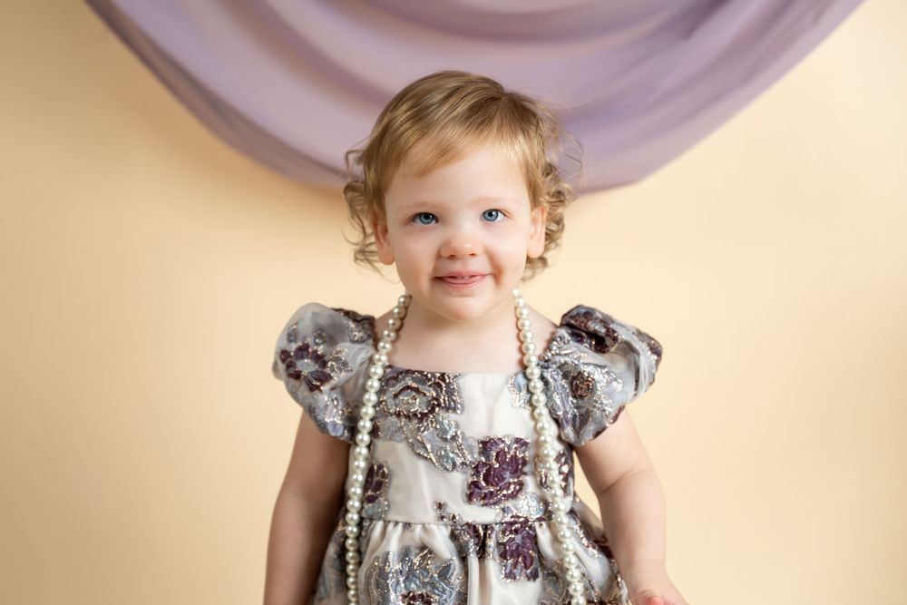 Smiling toddler girl in floral dress with pearl necklace poses against soft beige and lavender backdrop.
