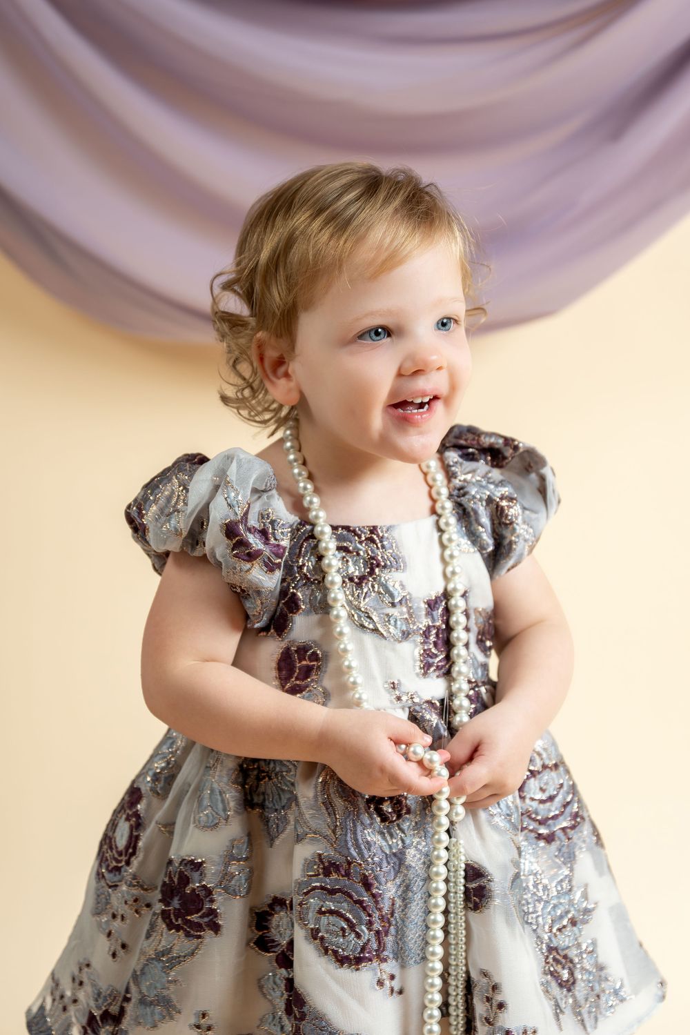 Smiling toddler girl in floral dress holding pearl necklace beads, studio portrait with purple drape backdrop.