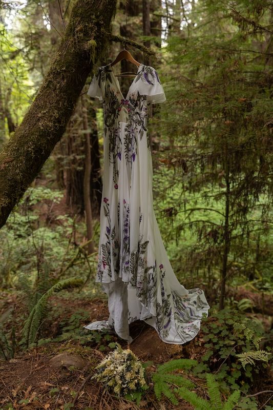 Floral wedding dress hanging on a wooden hanger from a mossy tree in a lush green forest setting.