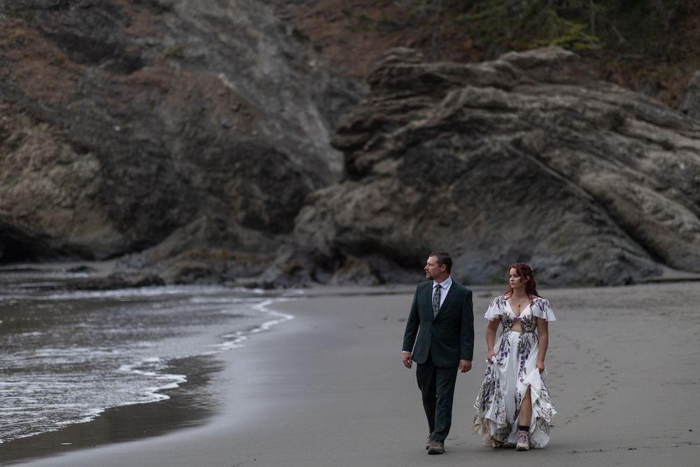 Couple in wedding attire walking hand in hand along a scenic rocky beach shoreline.