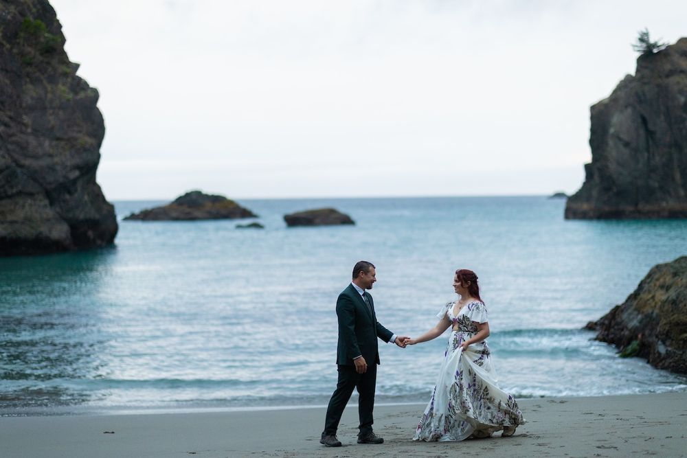Couple holding hands on a scenic beach with rocky sea stacks and turquoise water, bride in floral gown.